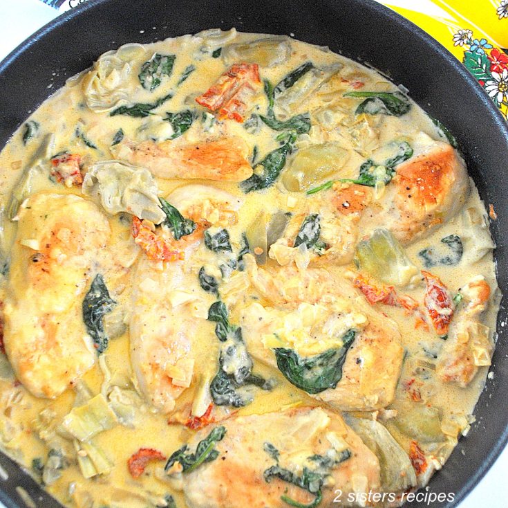 A black skillet with pieces of cooked chicken, artichokes, spinach and sundried tomatoes in a cream sauce.