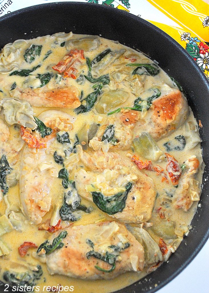 A large black skillet filled with pieces of chicken, spinach, artichokes smothered in a cream sauce.