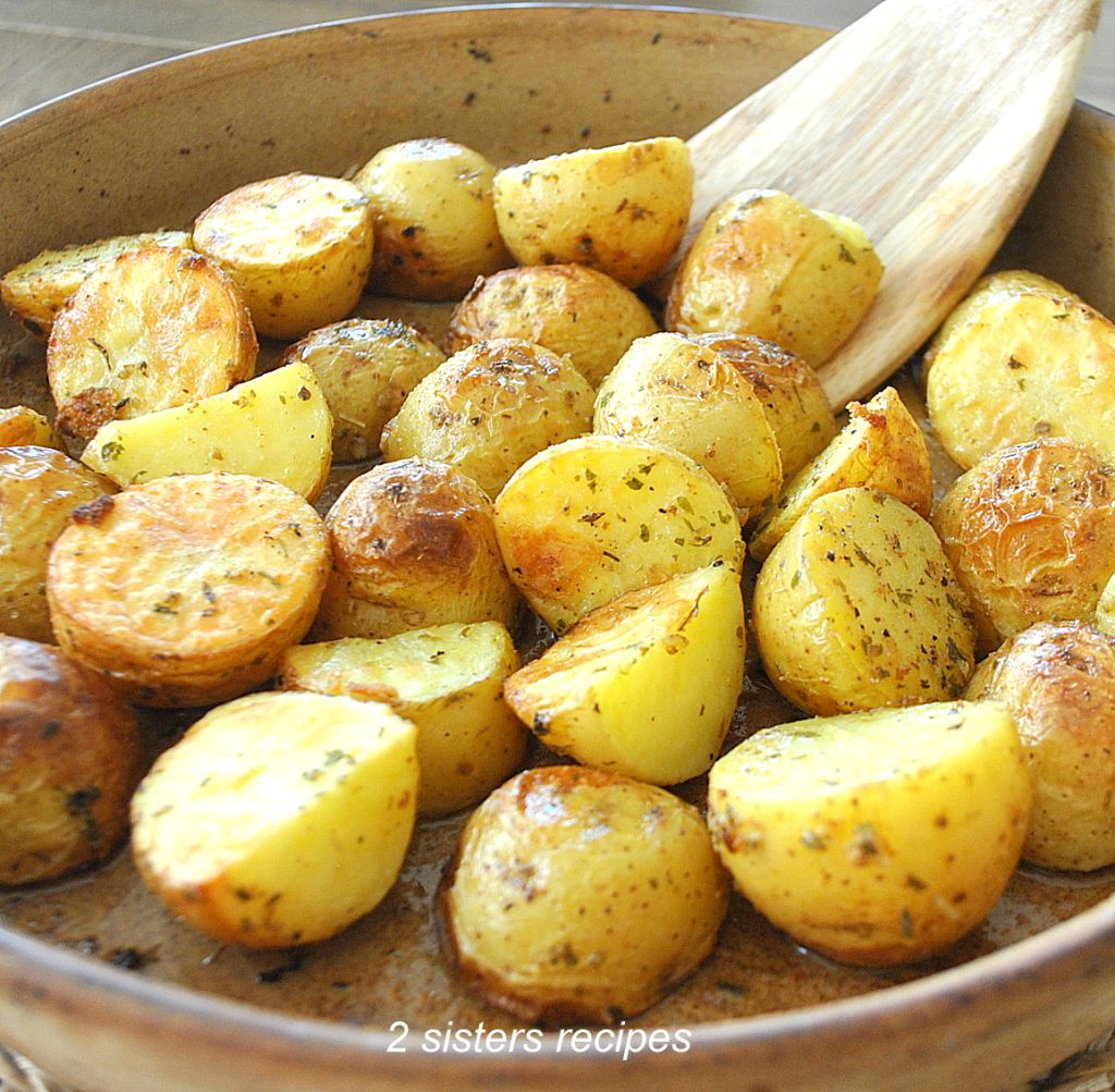 A wooden spoon is tossing the roasted potatoes in a brown baking dish. 