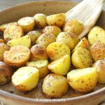 A wooden spoon is tossing the roasted potatoes in a brown baking dish.