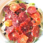A white round dish with slices of blood oranges over sliced fennel, capers and raisins.