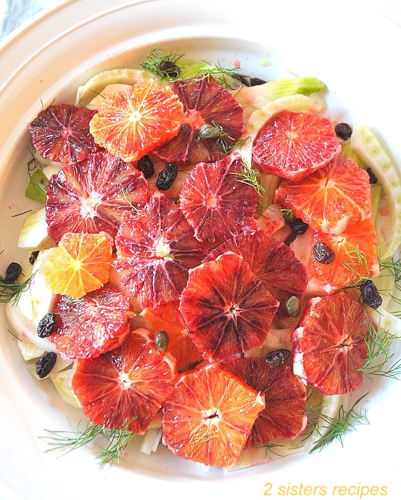 A white round dish with slices of blood oranges over sliced fennel, capers and raisins.