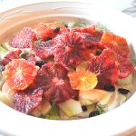 A white plate filled with sliced blood oranges over sliced fennel with capers and raisins.