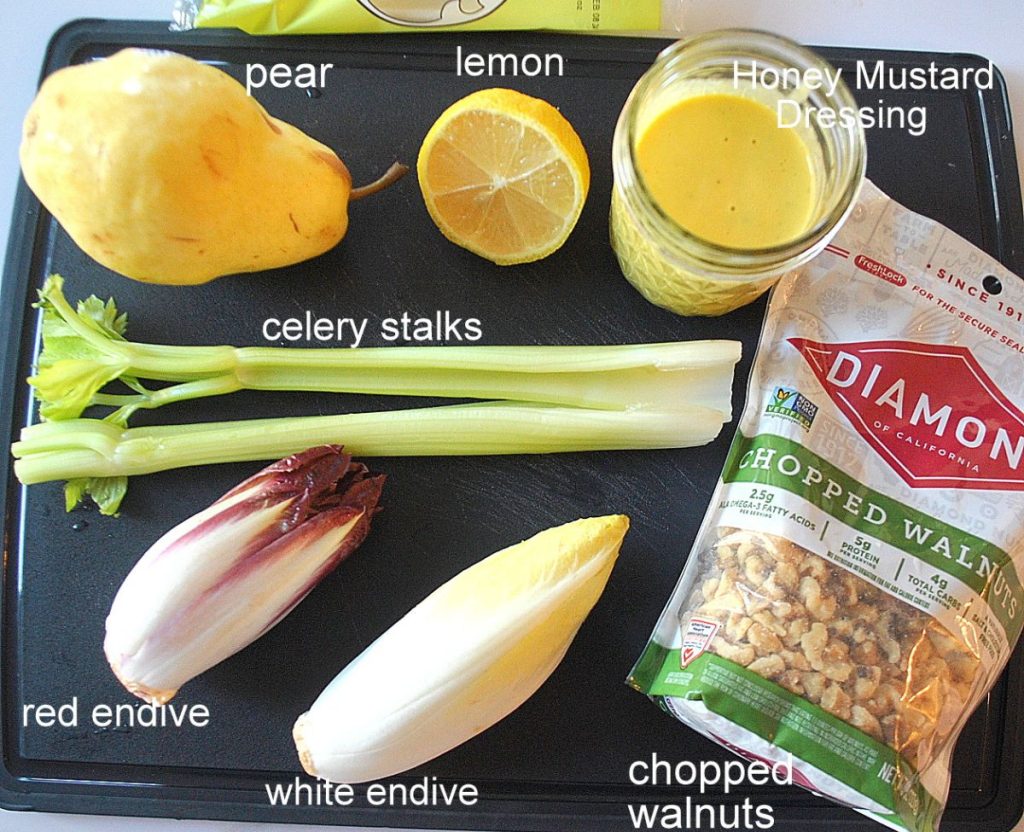 Ingredients displayed on a black board, one pear, lemon, jar of dressing, 2 bulbs of endives, stalks of celery and a package of chopped walnuts.