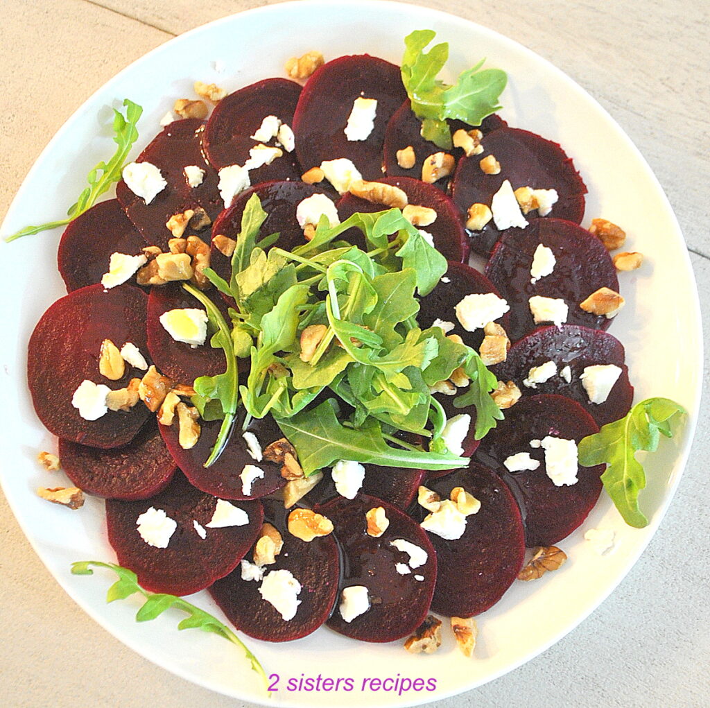 A white plate with sliced beets, topped with some arugula, chopped walnuts and crumbled goat cheese.