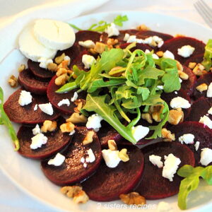 A white plate with sliced beets, topped with some arugula, chopped walnuts and crumbled goat cheese with 2 slices of goat cheese on the plate.