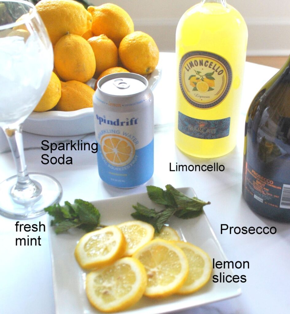 A bowl of lemons, a bottle of Limoncello, and Prosecco, can of sparkling soda, and sliced lemons on a white plate.
