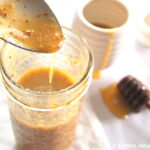 A spoonful of Dijon honey vinaigrette poured back into a mason jar. And a honey stick on the side on the table.