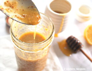 A spoonful of Dijon honey vinaigrette poured back into a mason jar. And a honey stick on the side on the table.