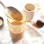 A spoonful of Dijon honey vinaigrette poured back into a mason jar. And a honey stick on the side on the table.