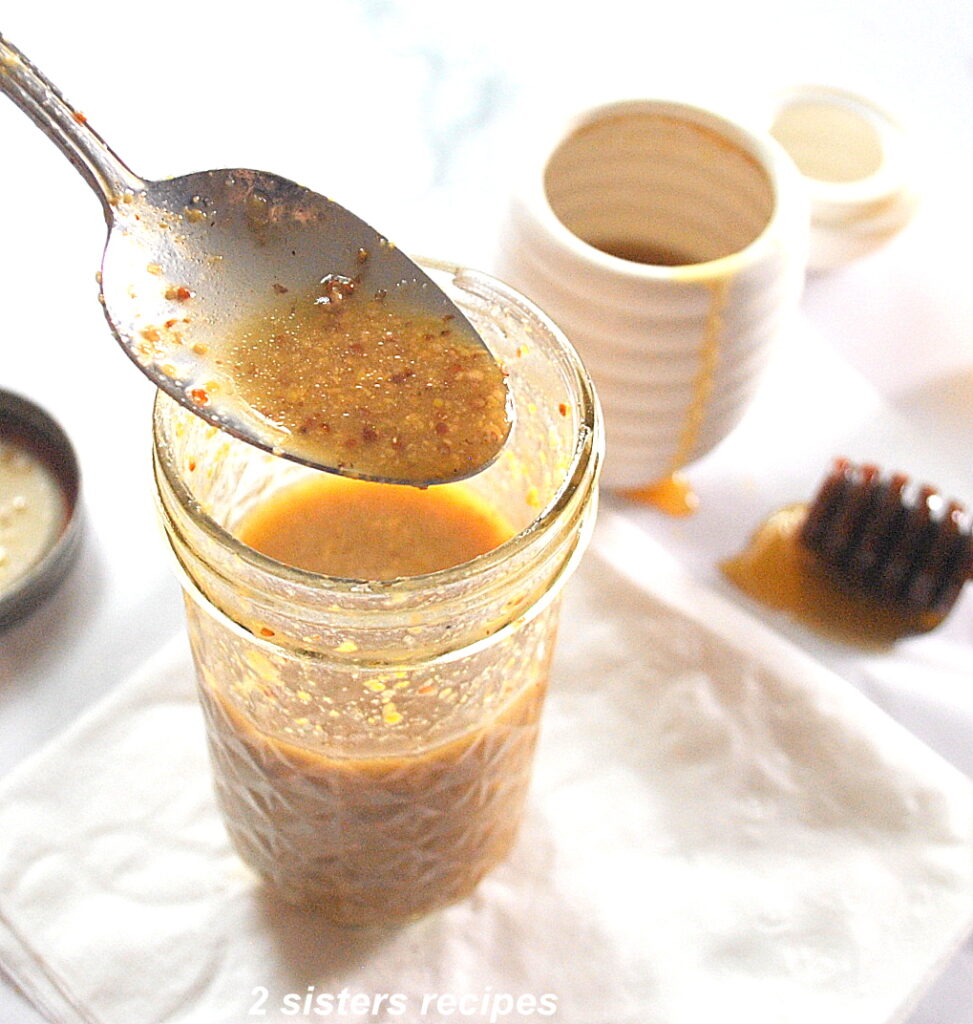 A spoonful of Dijon honey vinaigrette poured back into a mason jar. And a honey stick on the side on the table.