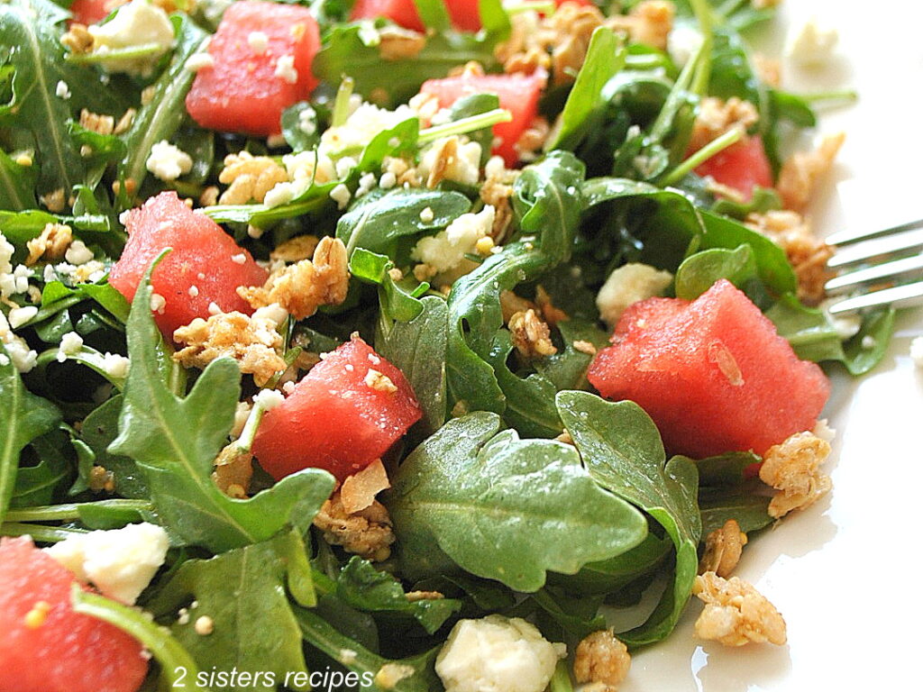On a white plate, a serving of salad loaded with melon bites, granola and crumbled cheese on top.