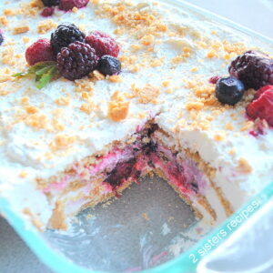 A glass baking dish with a white cake, topped with crushed crackers and mixed berries.