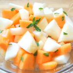 Cubes a white and orange melon tossed with fresh mint leaves in a salad bowl.