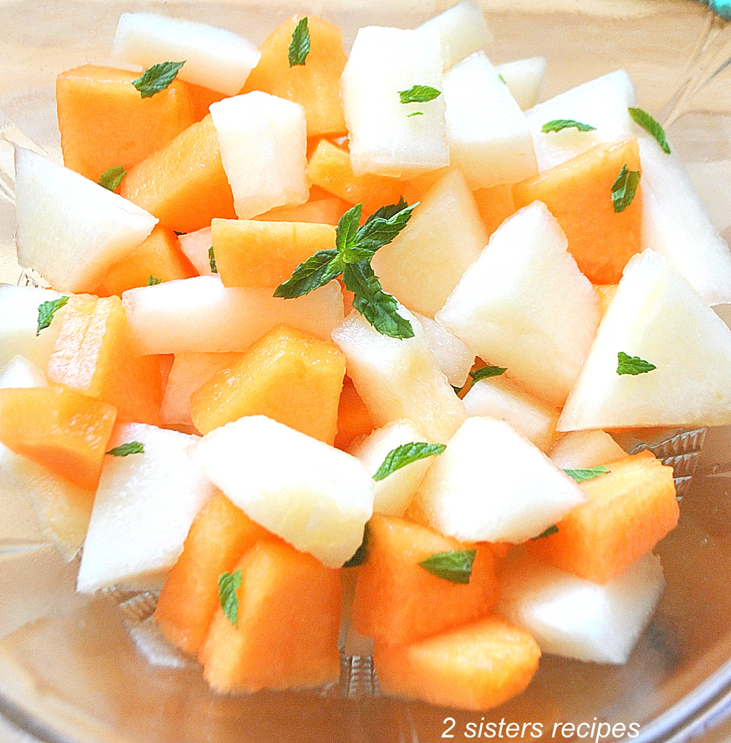 white and orange colored melons cut into pieces tossed in a clear bowl with mint leaves.