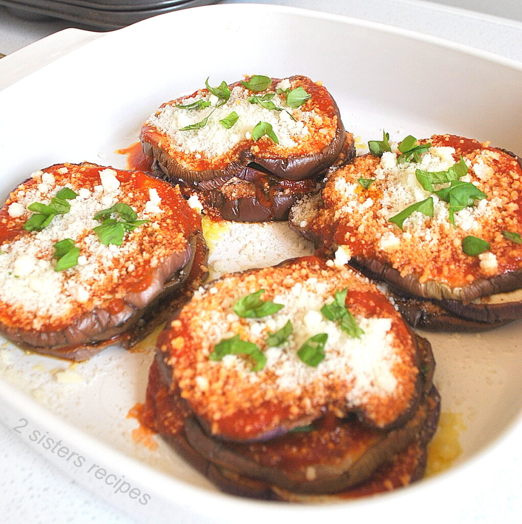 Four eggplant stacks baked in a white baking dish topped with tomato sauce, grated cheese and basil.