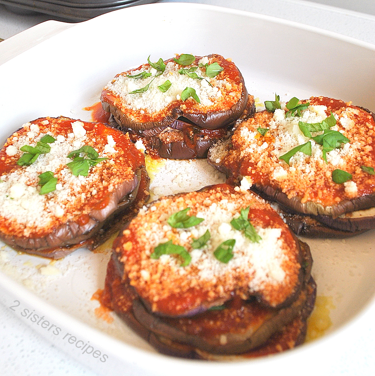 Four eggplant stacks baked in a white baking dish topped with tomato sauce, grated cheese and basil.