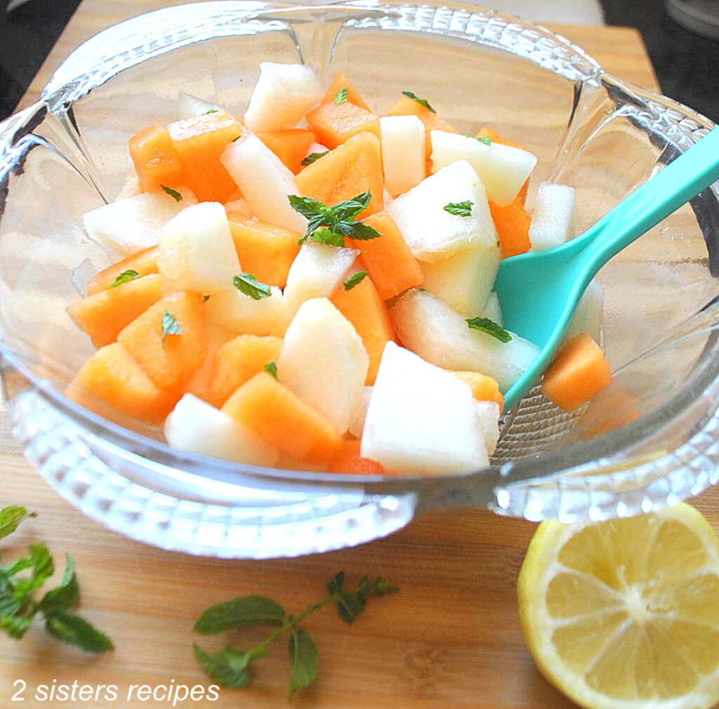 A blue ladle inside a clear salad bowl filled with white and orange melon pieces and mint.