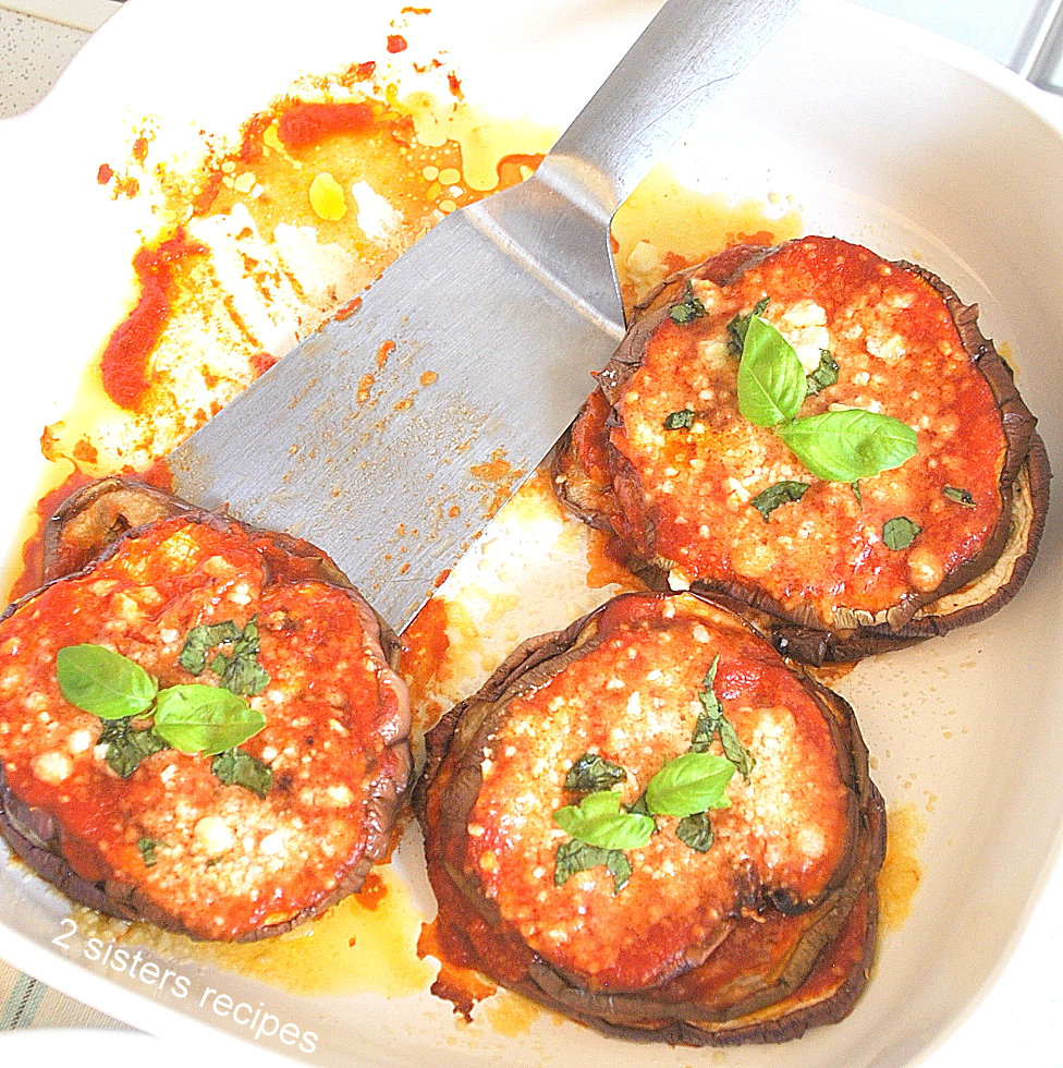 2 stacks of eggplants topped with tomato sauce, grated cheese and fresh basil in a white baking sheet with a silver spatula.