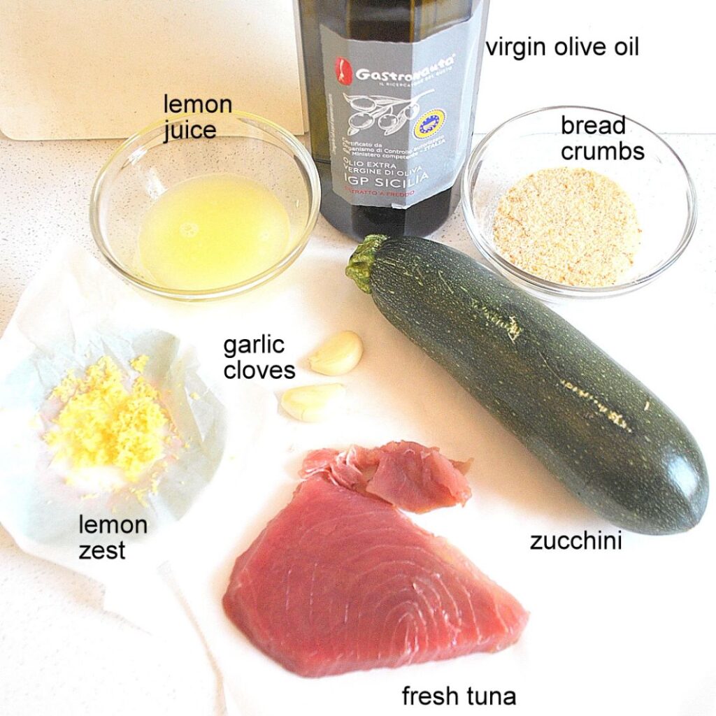 On parchment paper, fresh tuna steak, lemon juice, one zucchini, a bottle of olive oil, lemon zest and a glass container with bread crumbs and some garlic cloves.