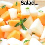 A clear bowl filled with cubes of white and orange melon with fresh mint leaves scattered on top.