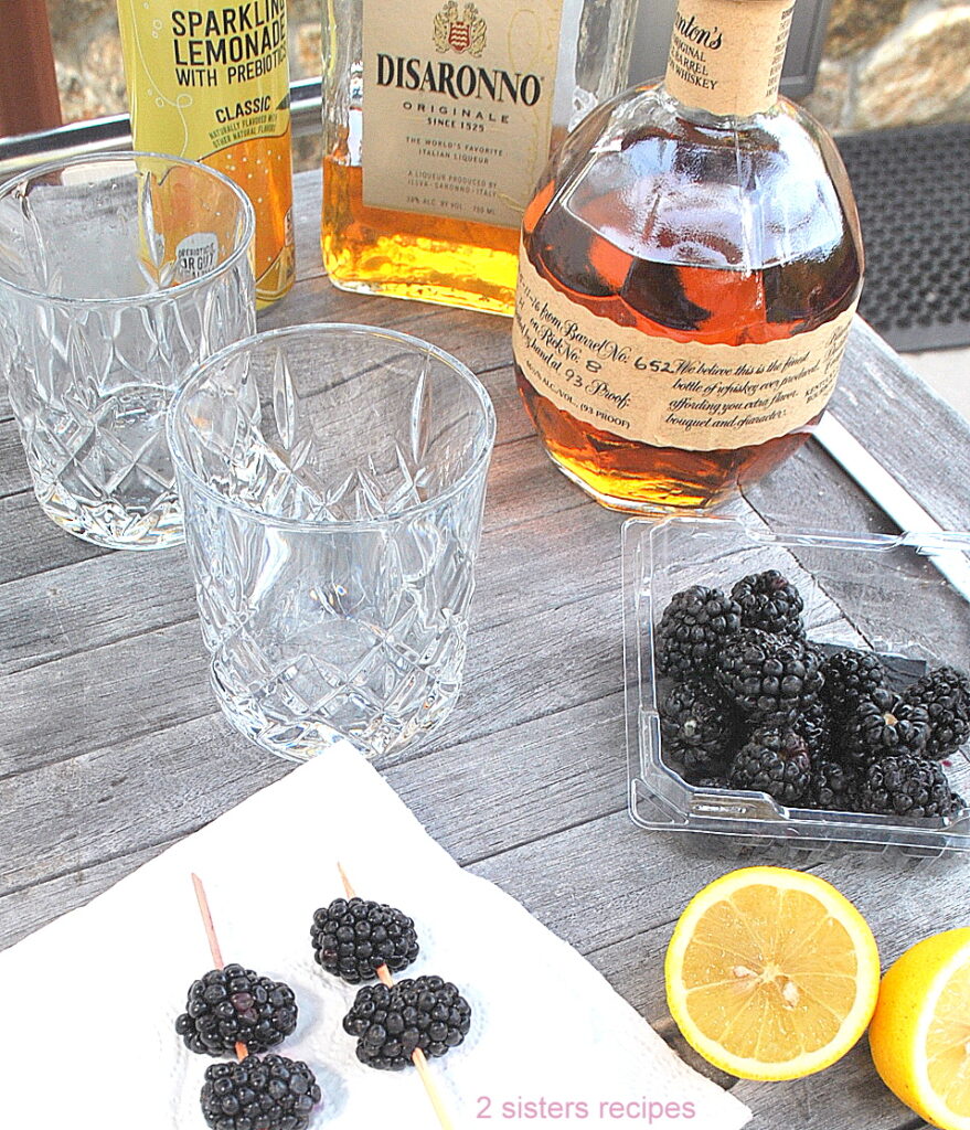 A bar with liqueurs, fresh blackberries, fresh lemon sliced in half, and 2 crystal glasses.