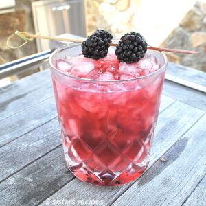 A crystal cocktail glass filled with a purple-pinkish color drink over crusted ice, topped with 2 fresh blackberries on a pick as garnish.