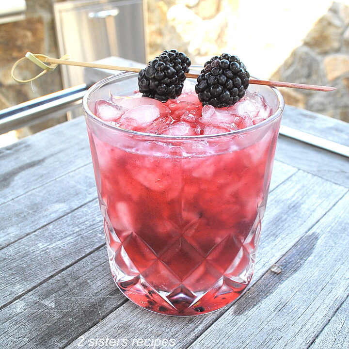 A crystal cocktail glass filled with a purple-pinkish color drink over crusted ice, topped with 2 fresh blackberries on a pick as garnish.