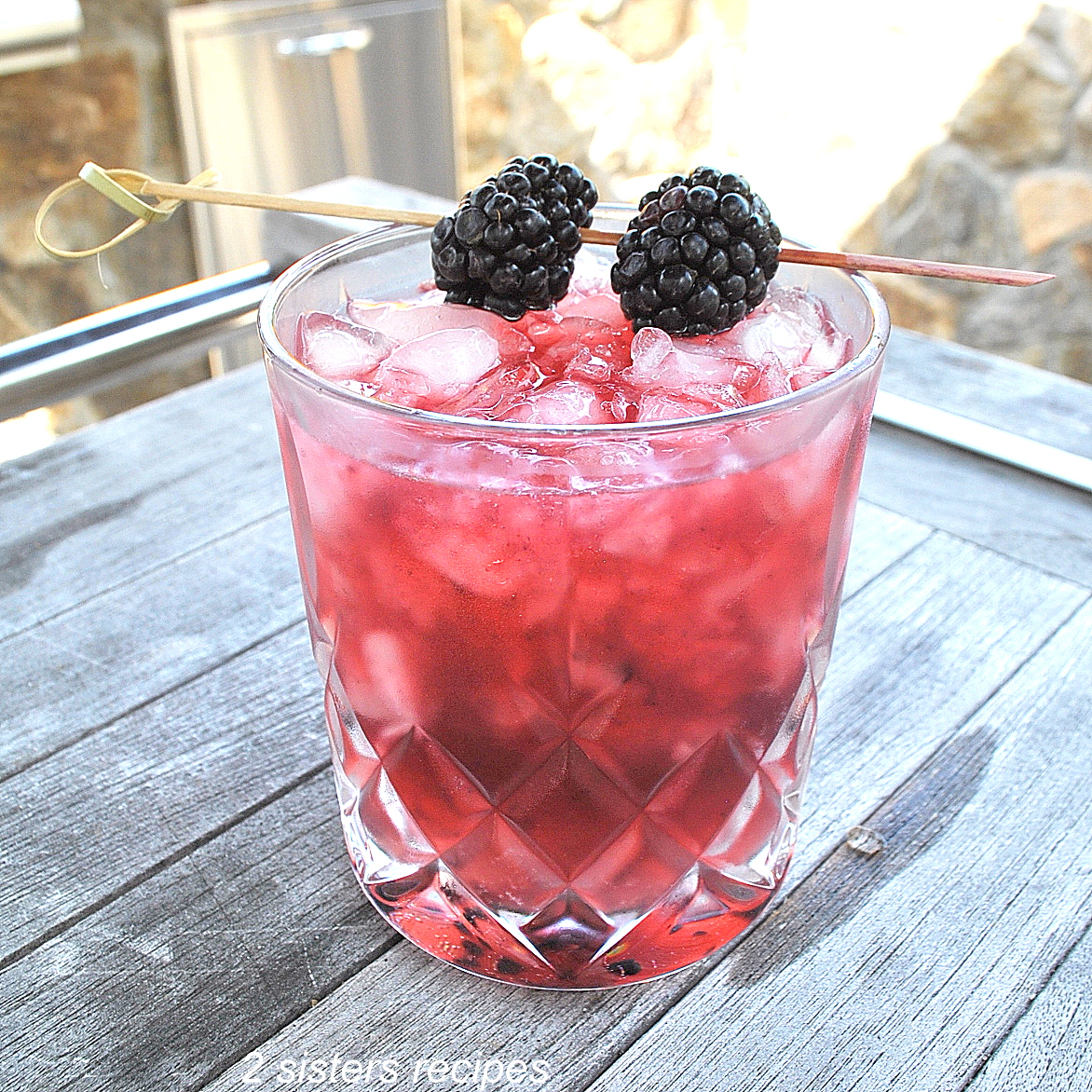 A crystal cocktail glass filled with a purple-pinkish color drink over crusted ice, topped with 2 fresh blackberries on a pick as garnish.