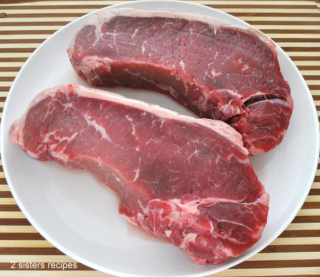 Two raw New York Strip steaks resting on a white plate.