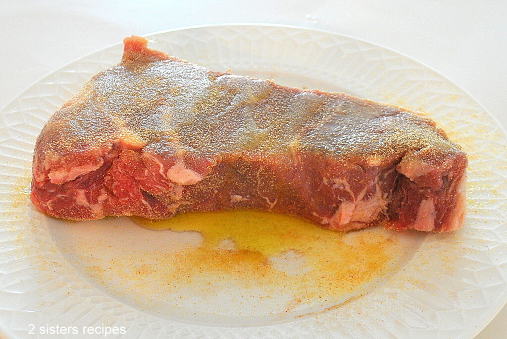 A raw steak on a white plate with garlic powder and olive oil drizzled on the steak.