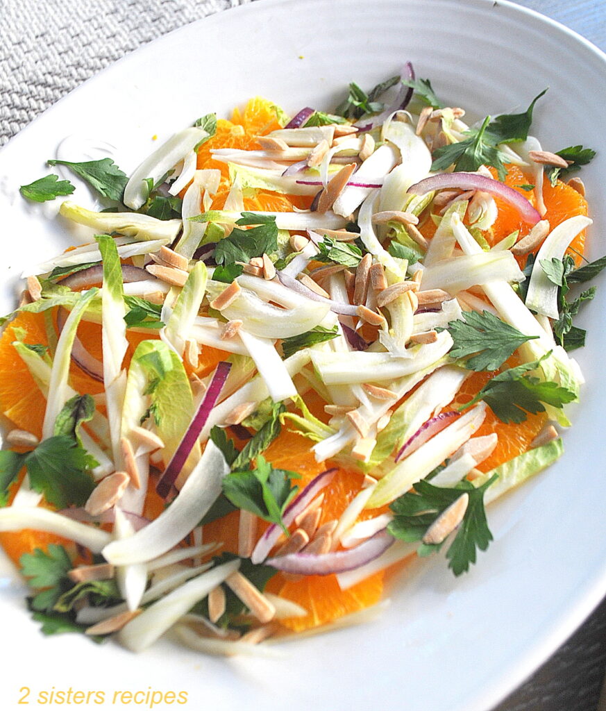 Am oval shape white platter filled with sliced endives, Navel oranges, red onion, and toasted almonds with parsley and mint leaves and served as a salad.