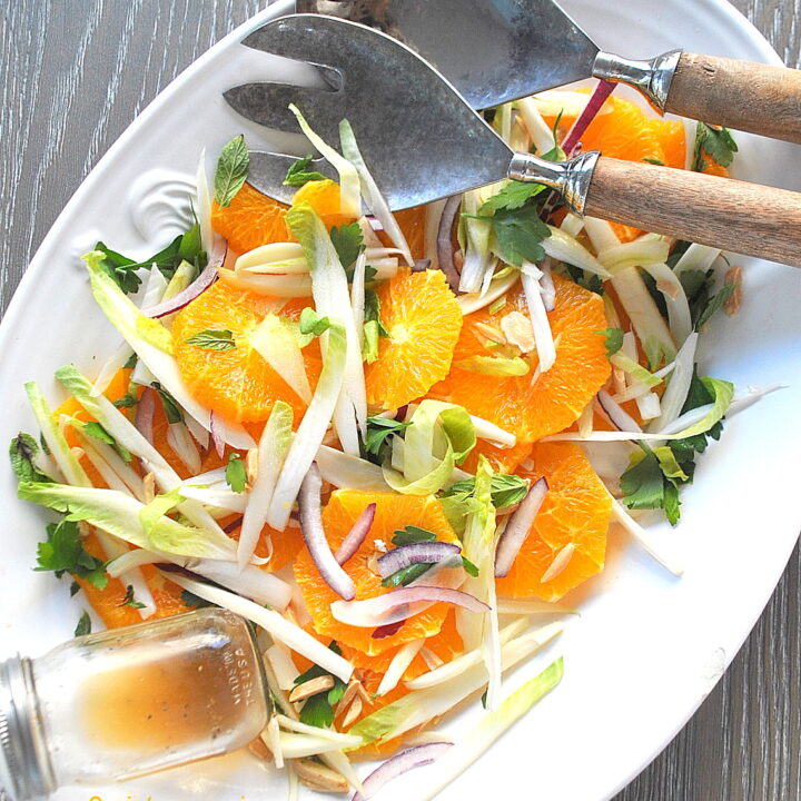 A white oval platter filled with sliced oranges, endives, red onions and tossed with fresh heabs.