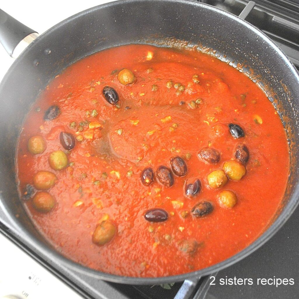 A black skillet on stovetop with a tomato sauce simmering with olives, and capers.