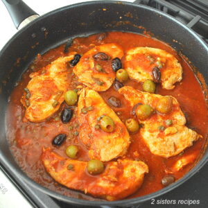 A black skillet filled with chicken smothered in a tomato sauce with olives and capers.