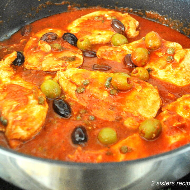 A large black skillet filled with Chicken breasts simmered in a puttanesca sauce with olives and capers.