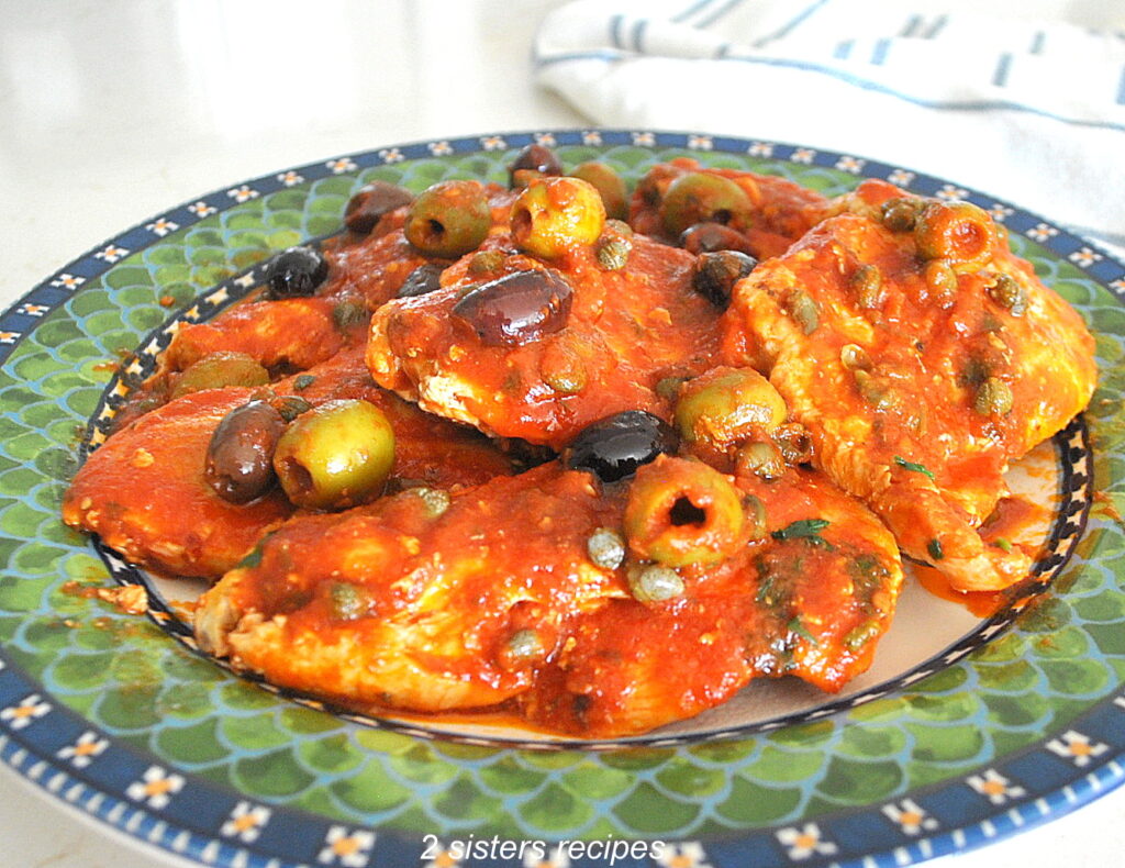 A green and blue colorful plate filled with pieces of chicken smothered in a puttanesca sauce,
