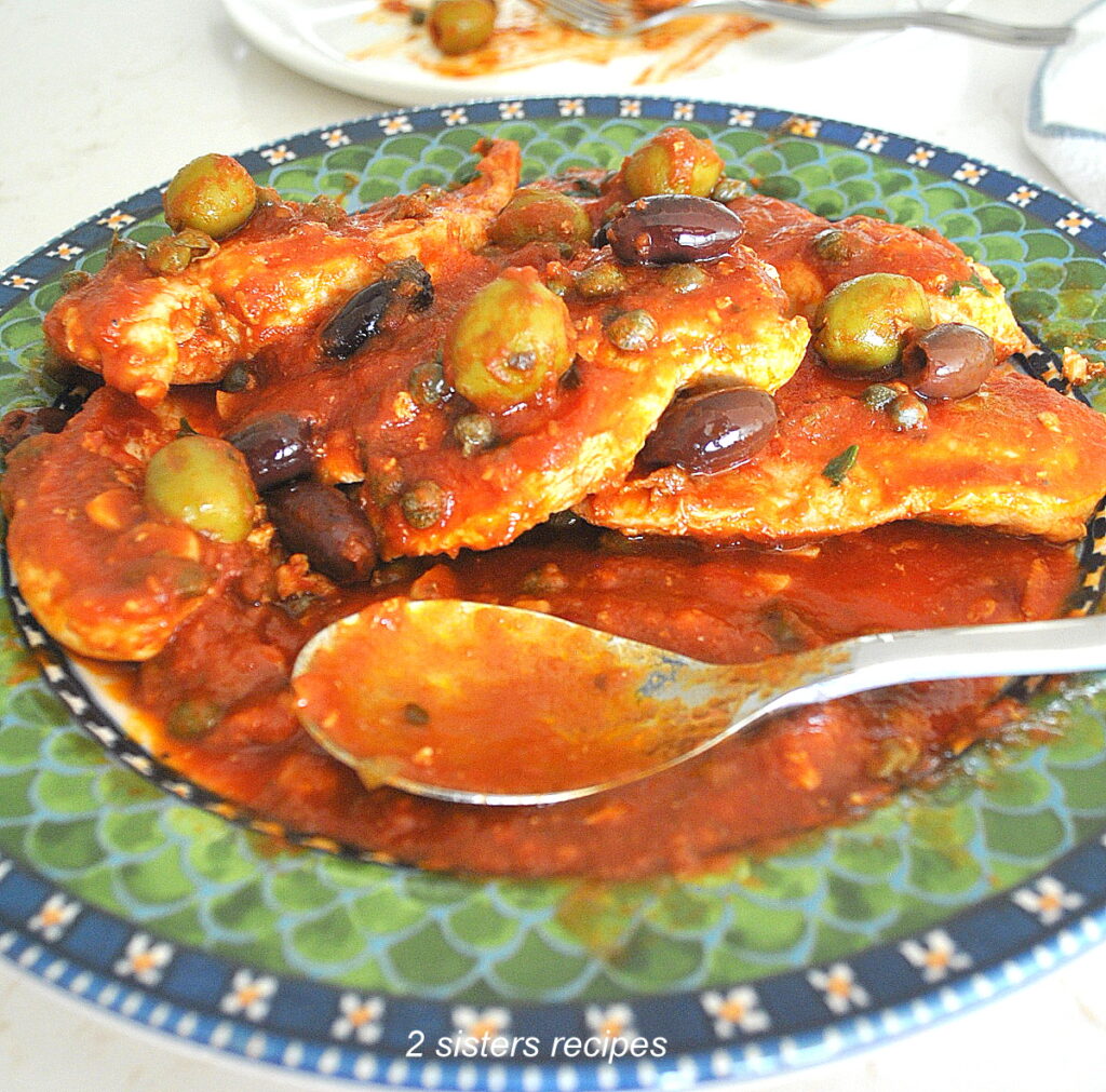 A green and blue dish filled with chicken pieces smothered in a puttanesca tomato sauce.