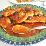 A green and blue dish filled with chicken pieces smothered in a puttanesca tomato sauce.