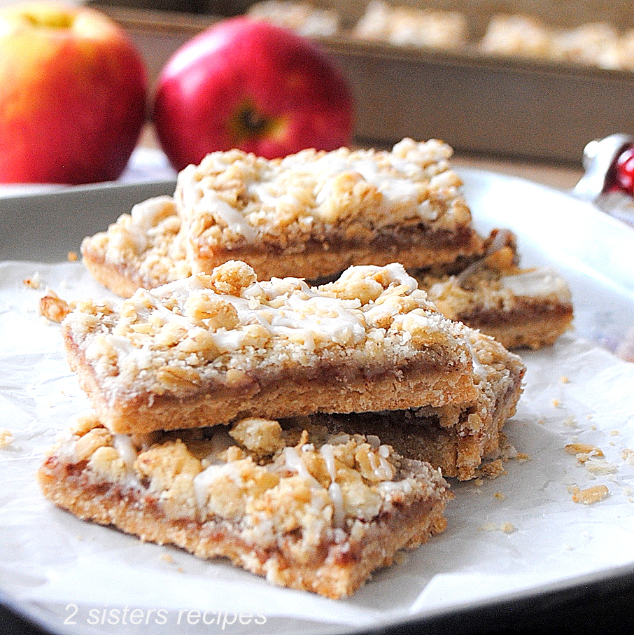 A pile of apple crisp bars set on a silver tray with 2 whole apples behind them.