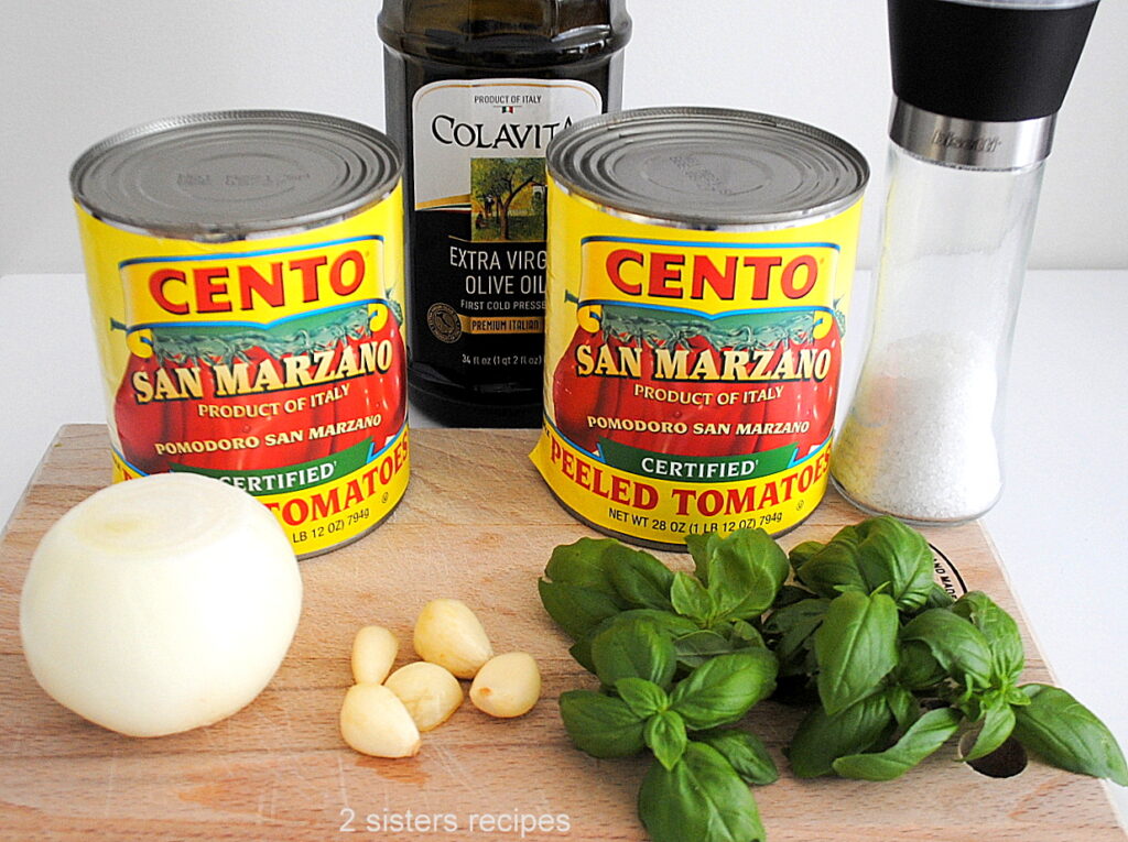 placed on a wood cutting board are 2 cans of San Marzano tomatoes, fresh basil, one onion, 5 cloves of garlic, bottle of olive oil and bottle of salt.
