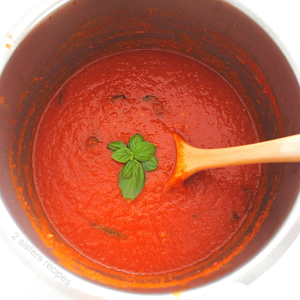 A large sauce pot filled with San Marzano Tomato sauce with fresh basil in the center, and a wooden spoon stirring.