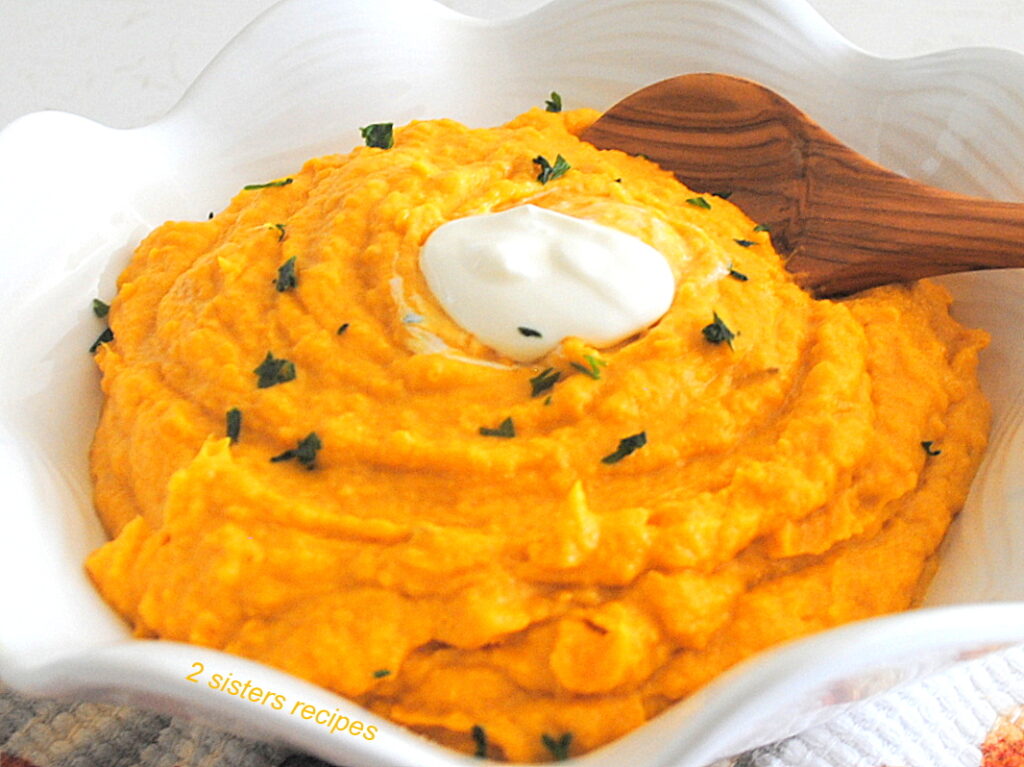 A white bowl filled with mashed pumpkin and sweet potatoes with a wooden spoon and a dollop of sour cream on top.