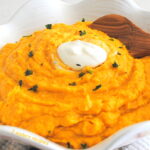 A white bowl filled with mashed pumpkin and sweet potatoes with a wooden spoon and a dollop of sour cream on top.