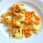 A white plate filled with ravioli topped with chunks of butternut squash and toasted pepitas.