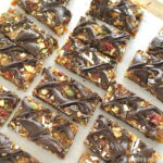 On sheet of parchment paper are fruit & nut protein bars with chocolate drizzled on top.