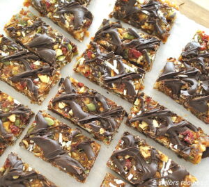 On sheet of parchment paper are fruit & nut protein bars with chocolate drizzled on top.