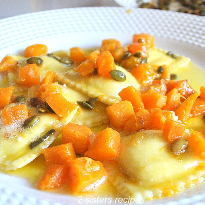 A white dinner plate filled with ravioli topped with chunks of butternut squash and toasted pepitas in a butter sauce.