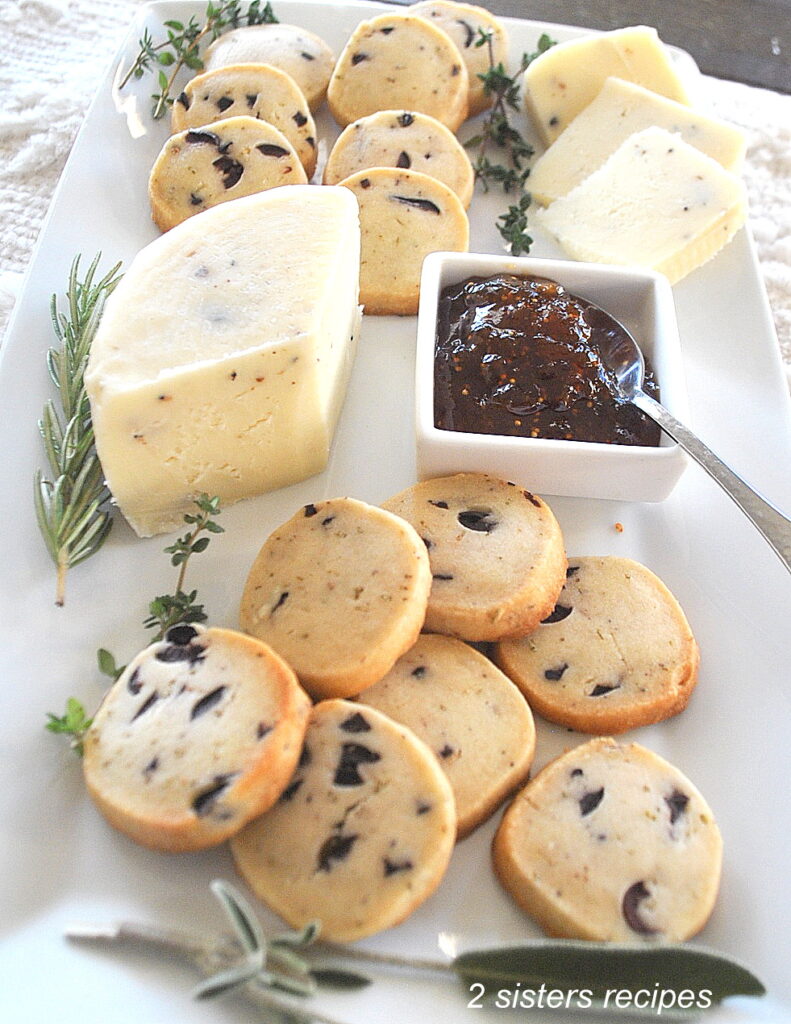 A white serving platter filled with olive rosemary biscuits, wedge of cheese, some jam with spoon, and fresh herbs as garnish.