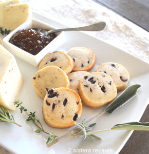 A white platter filled with shortbread biscuits studded with black olives, served with fig jam as an appetizer.